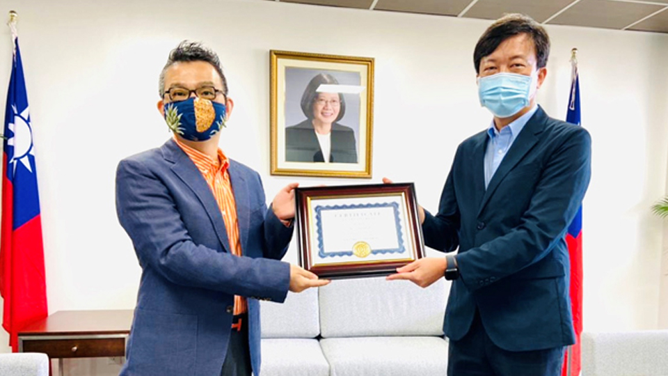Edward Pingyuan Lu stands with another person, they are holding a certificate. In the background there are two flags and a photograph of a person on the wall. 